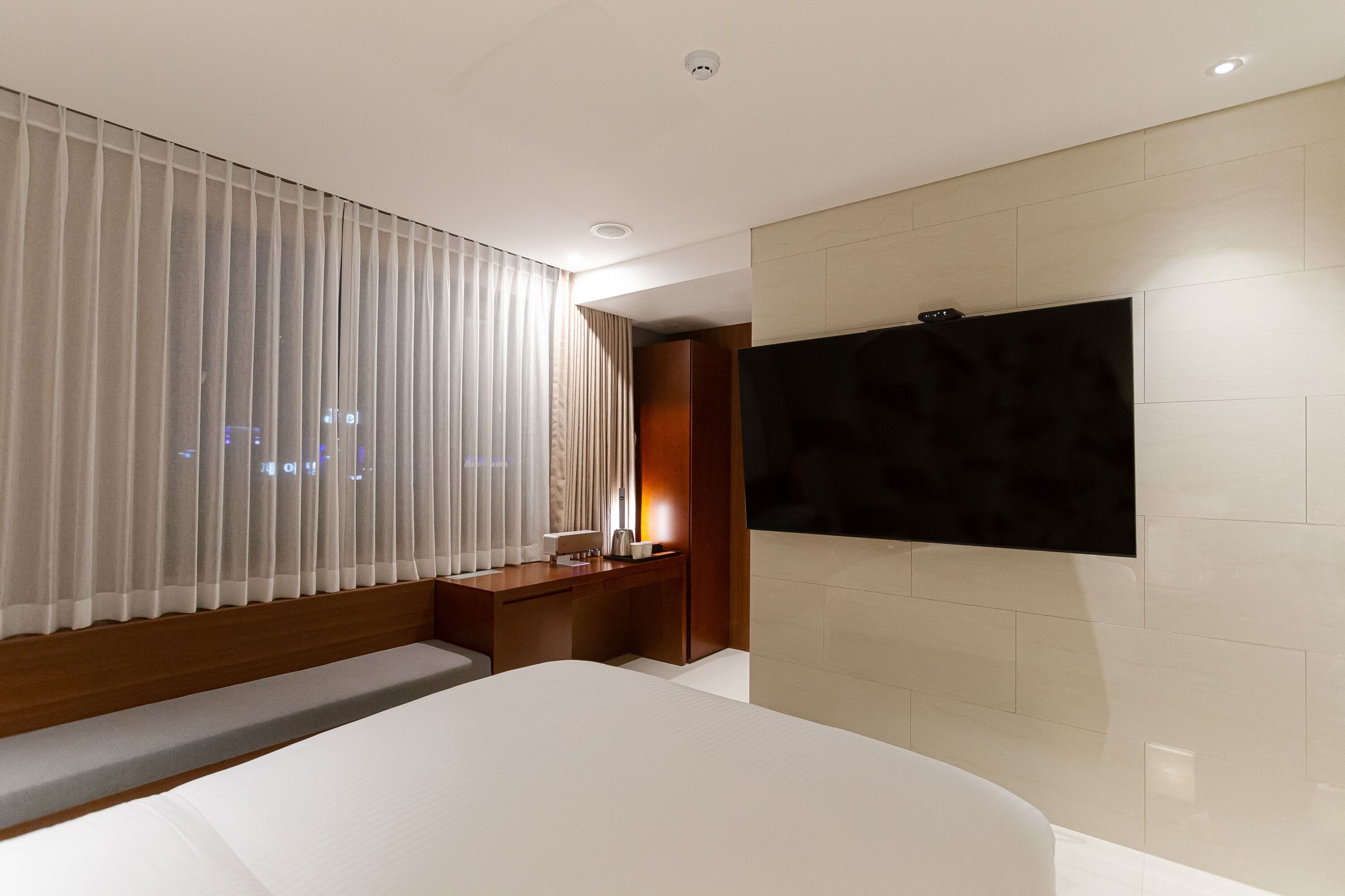 A modern hotel room with a large bed, a bedside table, a TV mounted on the wall, and a window with curtains.