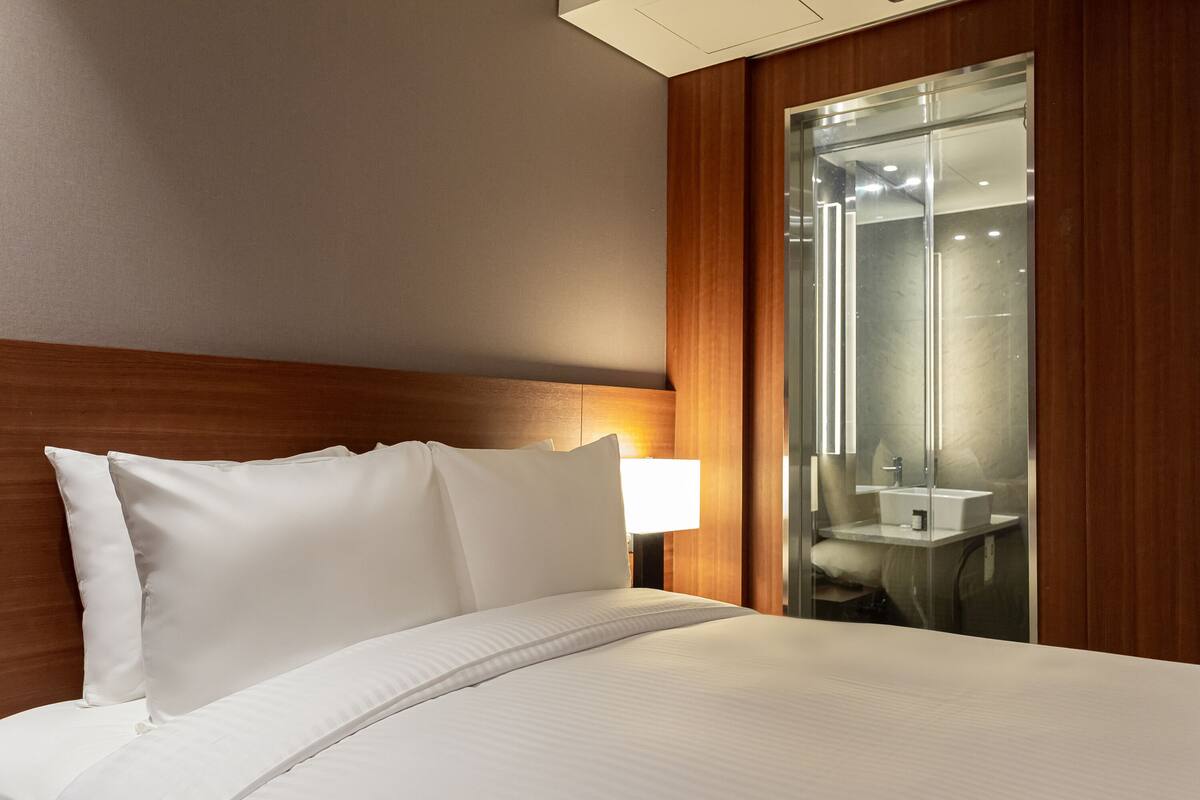Ein Hotelzimmer mit einem ordentlich bezogenen Bett, einem Holzkopfteil, einer Nachttischlampe und einem durch eine Glaswand abgetrennten Badezimmer mit Waschbecken und Spiegel.