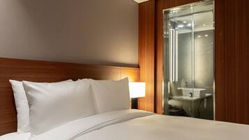 A hotel room with a neatly made bed, a wooden headboard, a bedside lamp, and a glass-enclosed bathroom with a sink and mirror.