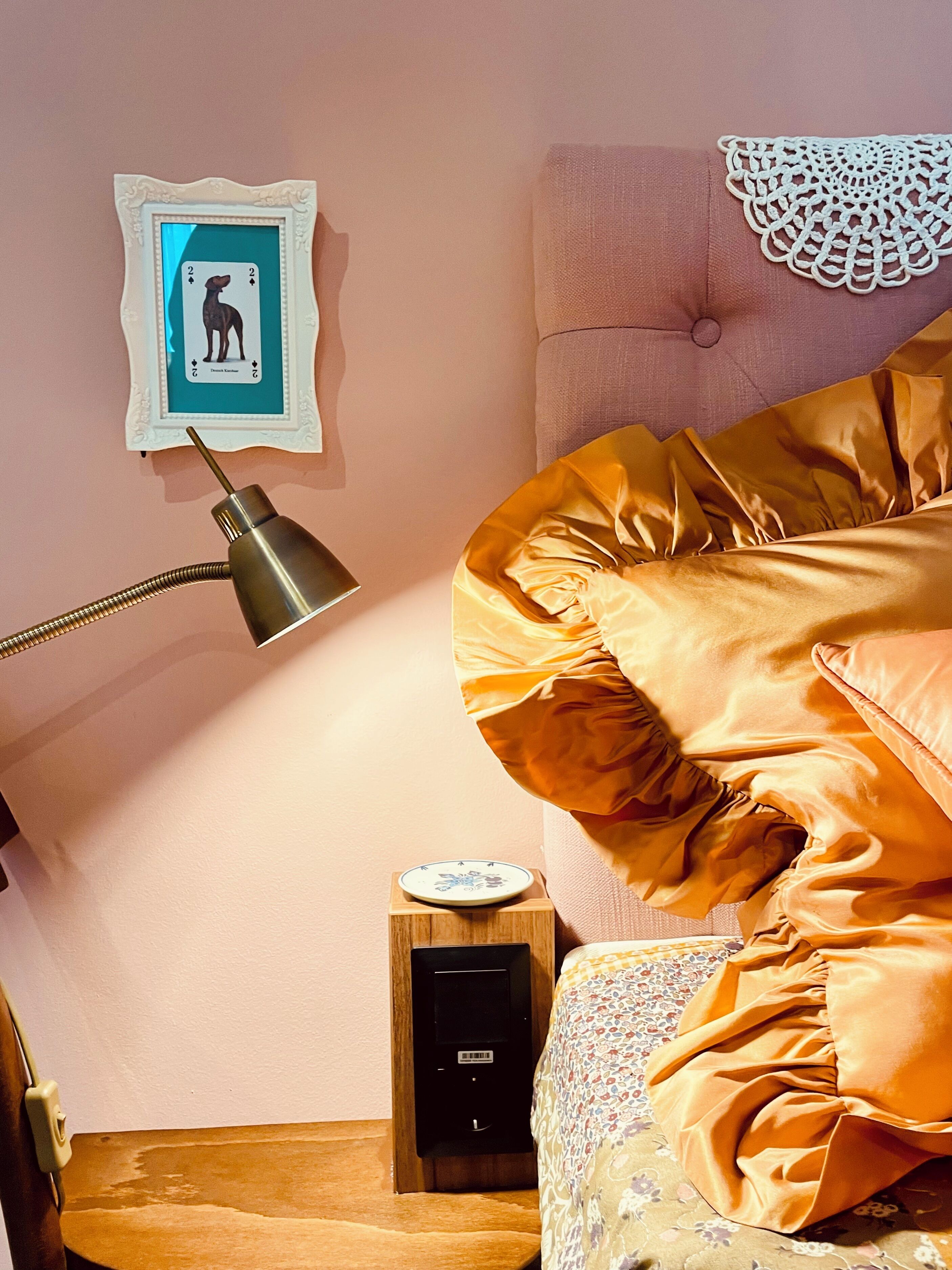 A bedroom with a bed, a bedside table with a lamp, a small bedside table with a plate, and a wall-mounted picture frame.