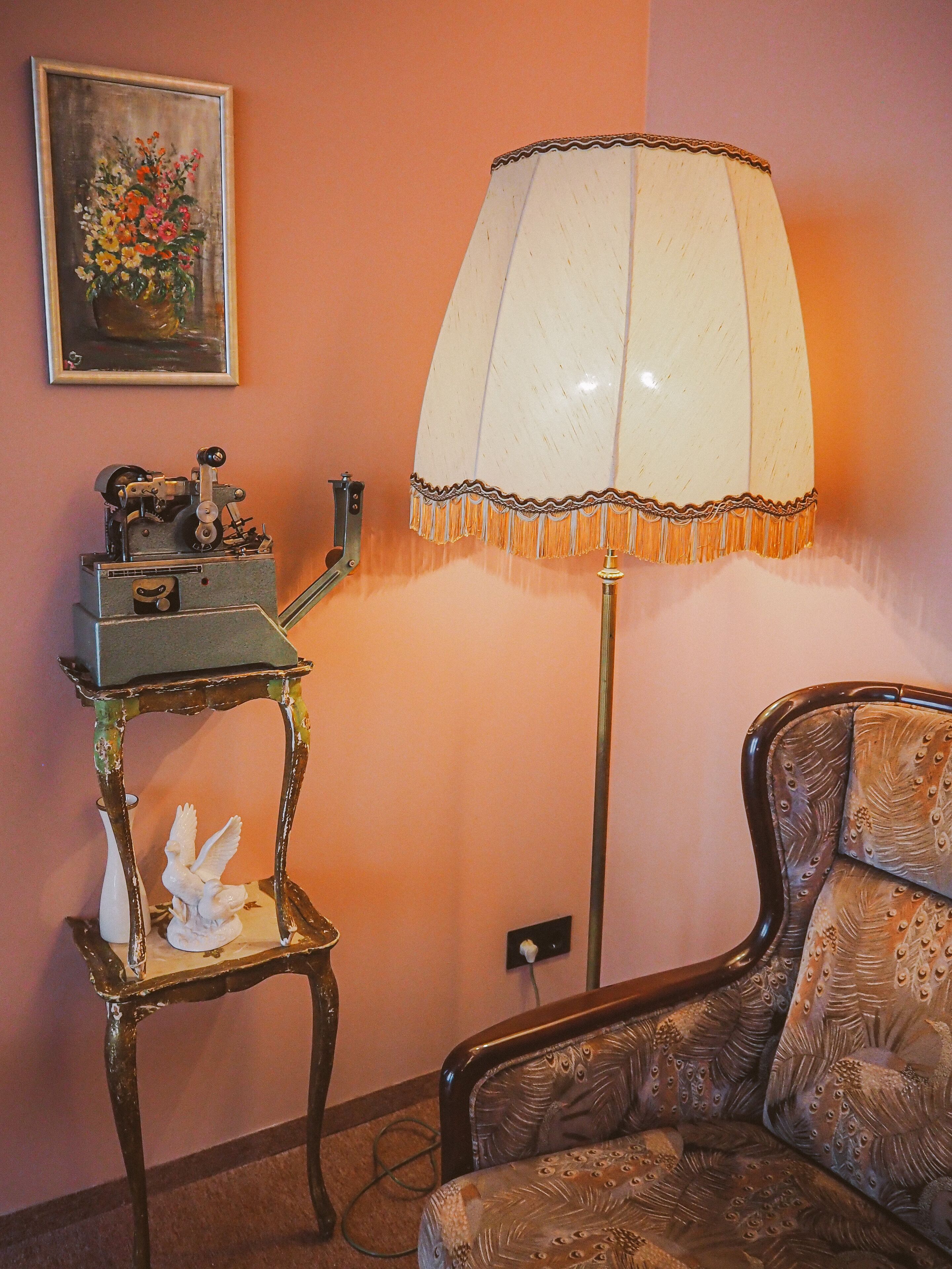 A room with a vintage-style lamp, a decorative table with a bird sculpture, and a chair with a peacock pattern.