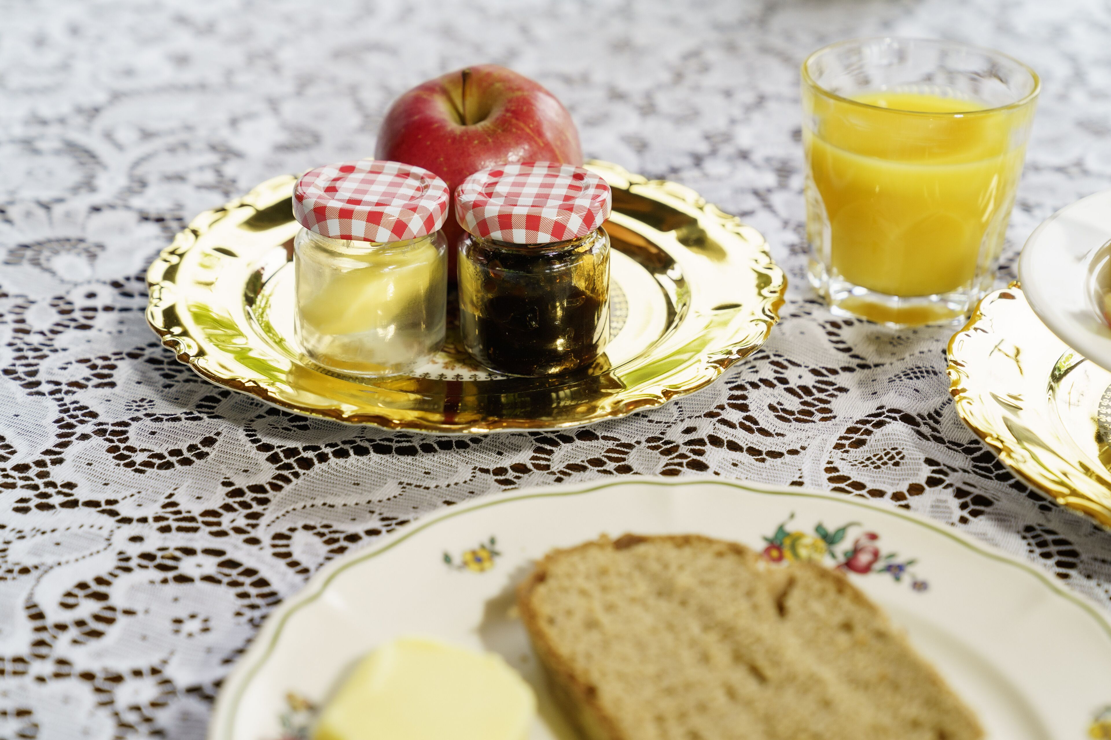 A table with a lace tablecloth, a plate with butter, a slice of bread, two jars with checkered lids, a glass of orange juice, and an apple.