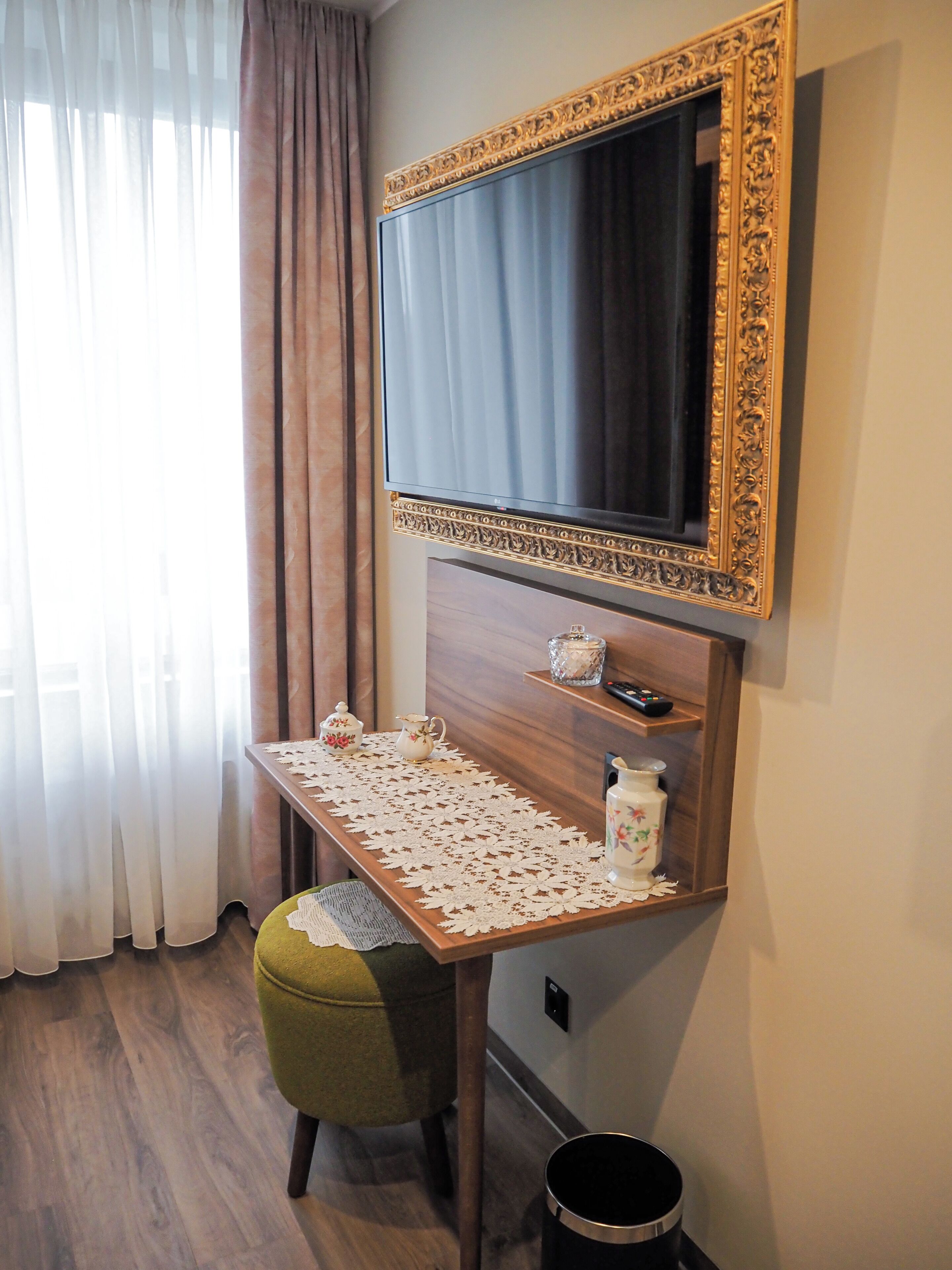 Una habitación con una televisión montada en una pared de madera, una mesita con mantel de encaje, una silla verde y un cubo de basura.