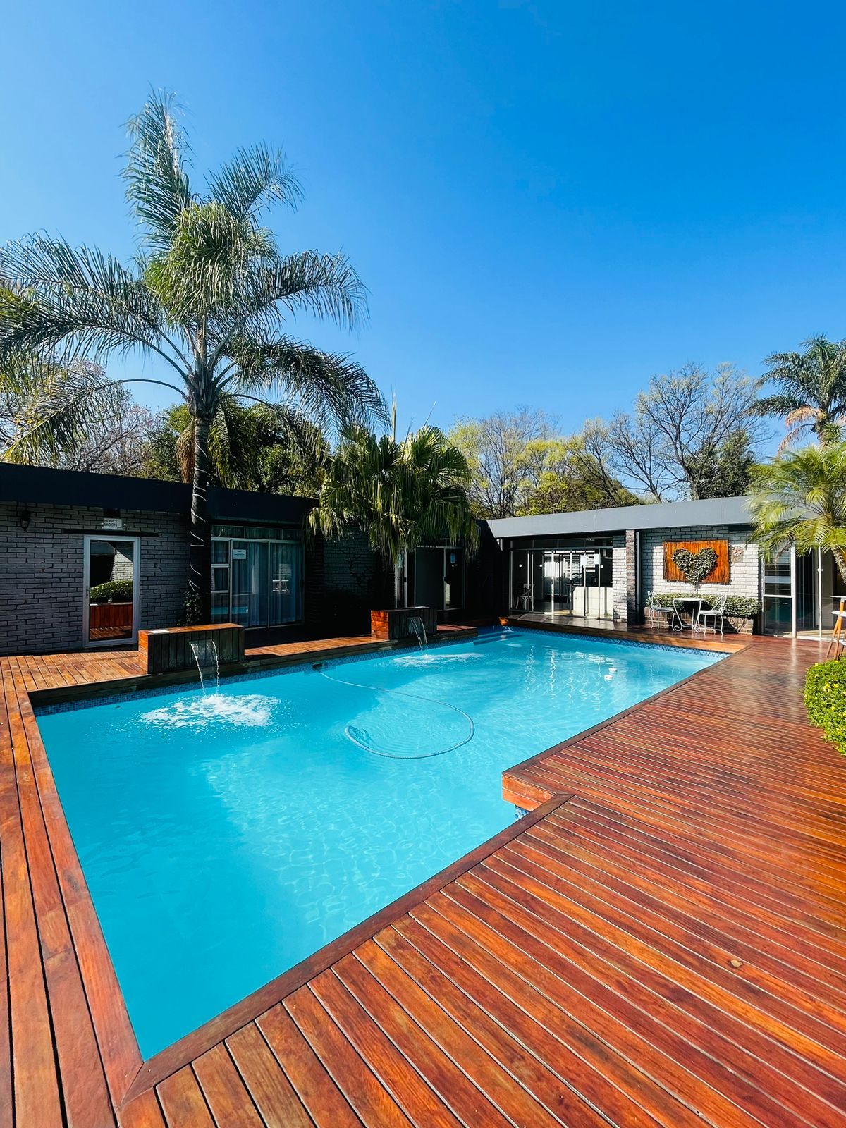 A swimming pool with a wooden deck, surrounded by buildings with brick and stone facades, and palm trees.