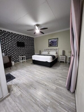 Un dormitorio con cama, ventilador de techo, pared con diseño de ladrillo, televisor montado y un cuadro en la pared.