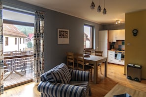 Uma sala de estar aconchegante com sofá, mesa de jantar e cadeiras. Uma varanda com deck de madeira e vista para casas é visível pela janela.
