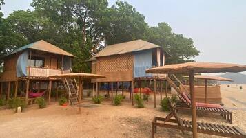 Un hébergement en bord de mer composé de deux cabanes en bois, chacune dotée d’un toit de chaume et d’un petit balcon. On y trouve également des chaises longues en bois et un toboggan pour la plage.