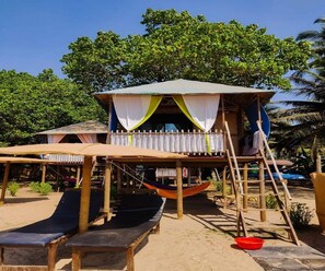 A beachside hut with a thatched roof, a hammock, and a small staircase leading to a platform with a white canopy.