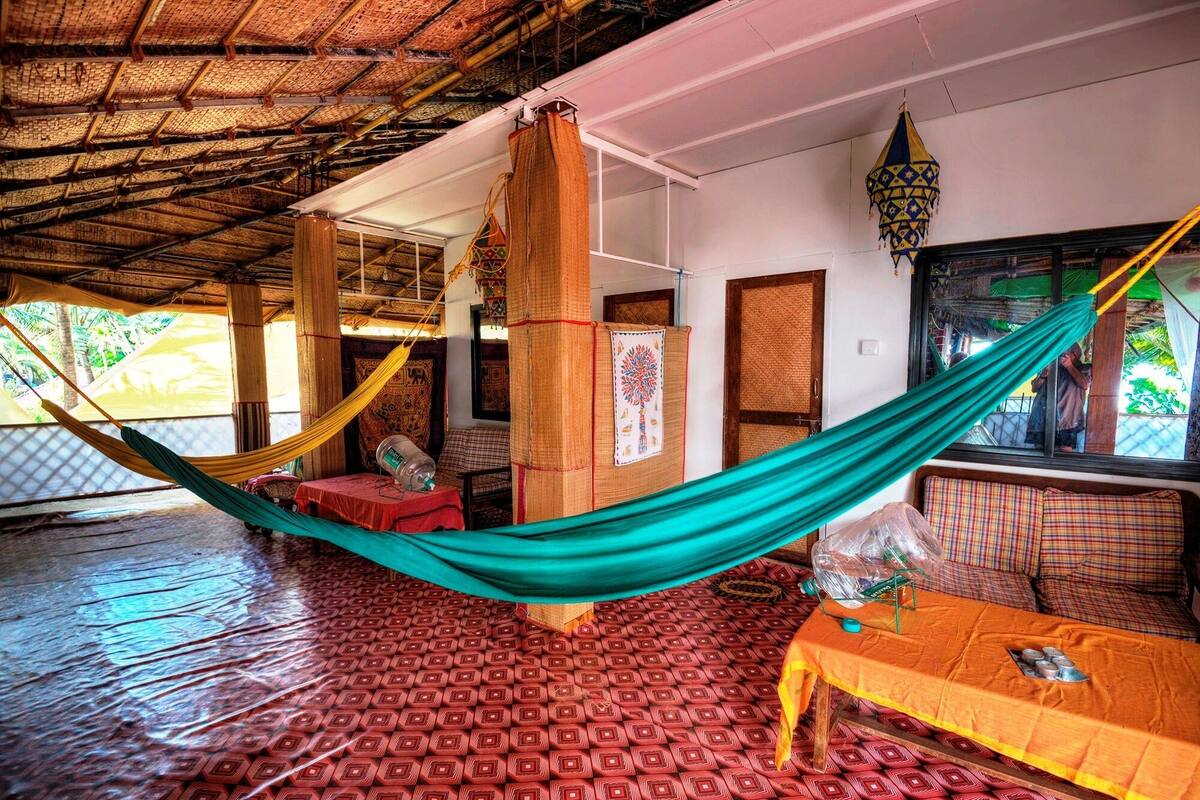 A hammock suspended between wooden pillars in a room with patterned flooring, a table with a yellow tablecloth, and a window with a view of greenery.