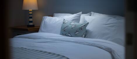 Een net opgemaakt bed met witte lakens en een gestreept dekbed, een kussen met een vogelprint, en een bedlampje op een houten nachtkastje.