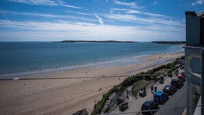 Une vue sur la plage, avec un ciel dégagé, une plage de sable et un bâtiment où sont garées des voitures, le tout entouré d’une clôture.