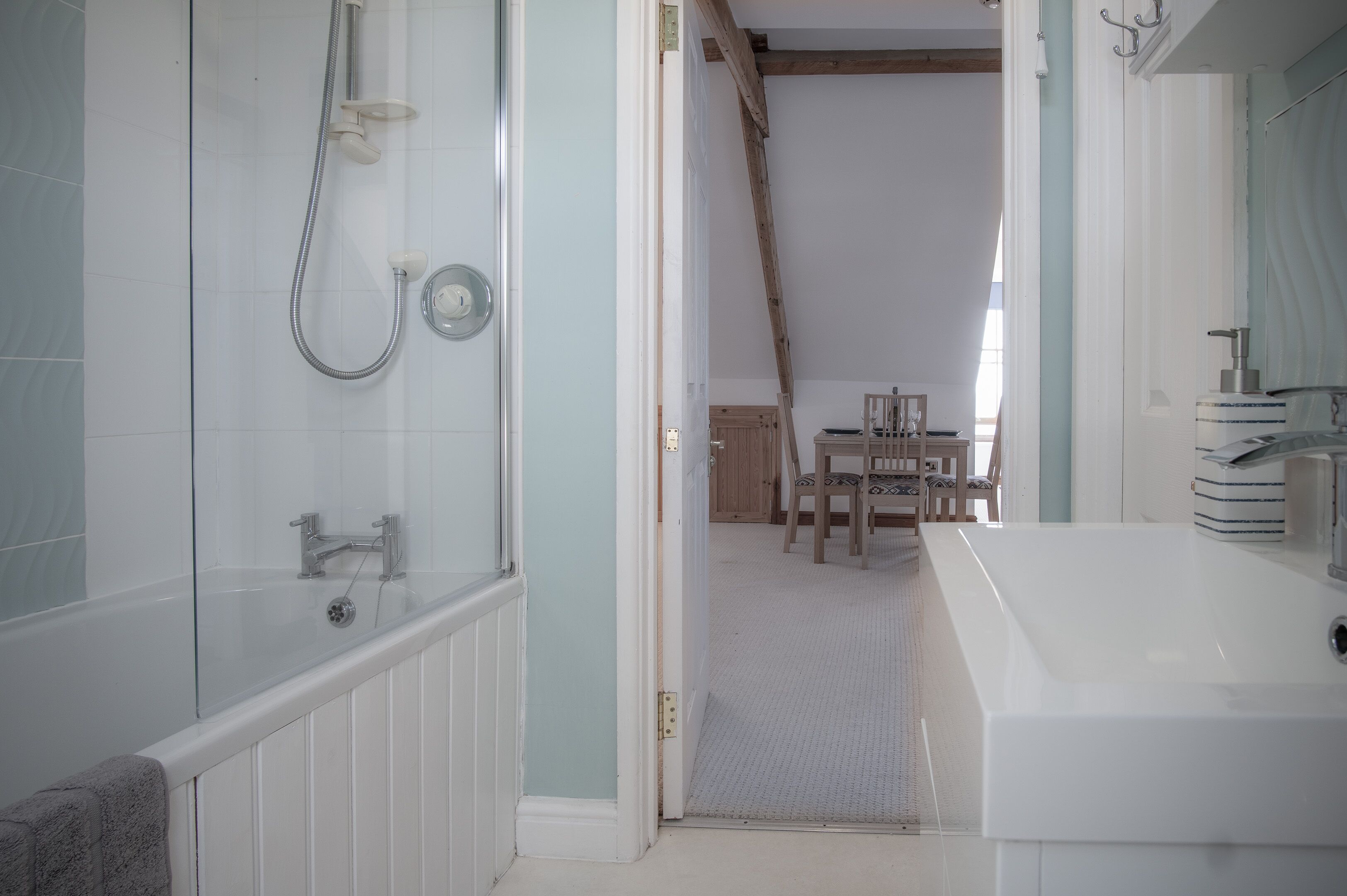 Ein Badezimmer mit Badewanne, Dusche und Waschbecken. Im angrenzenden Raum befindet sich ein Holzstuhl und ein Tisch.