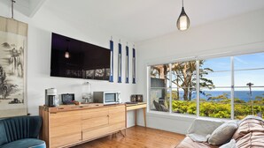 A modern living room with a wooden desk, a blue armchair, a brown sofa, and a large window offering a view of the sea. - Romantic Ocean View Studio @ Stanwell Tops (Stanwell Tops)