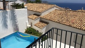 A rooftop pool with a clear view of the ocean, a white building with terracotta roof tiles, and a palm tree.