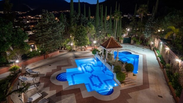 A pool area with a geometric tile design, surrounded by lounge chairs and a small building with a red roof.