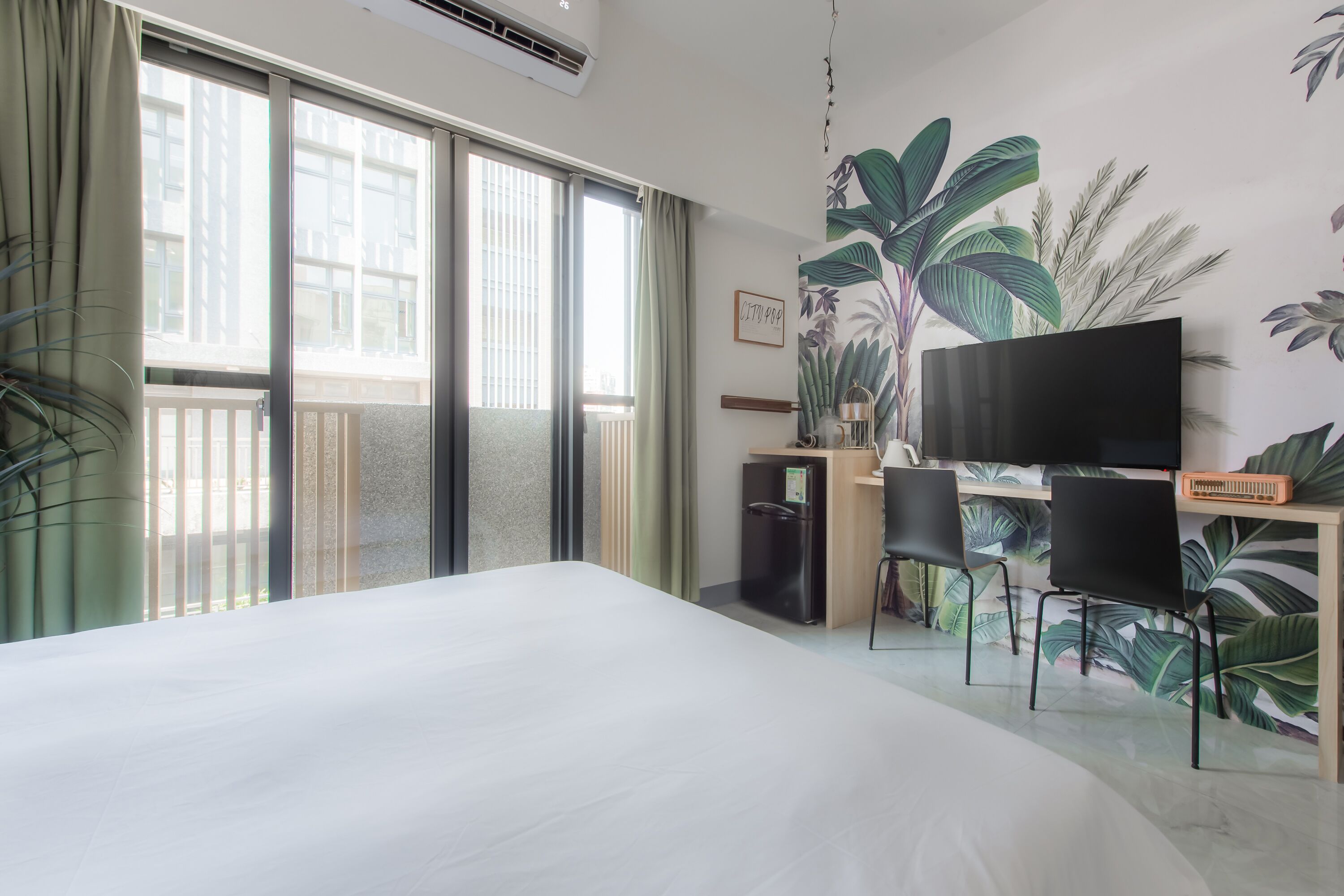 A hotel room with a large bed, a desk with a TV, and a balcony with a view of the city.
