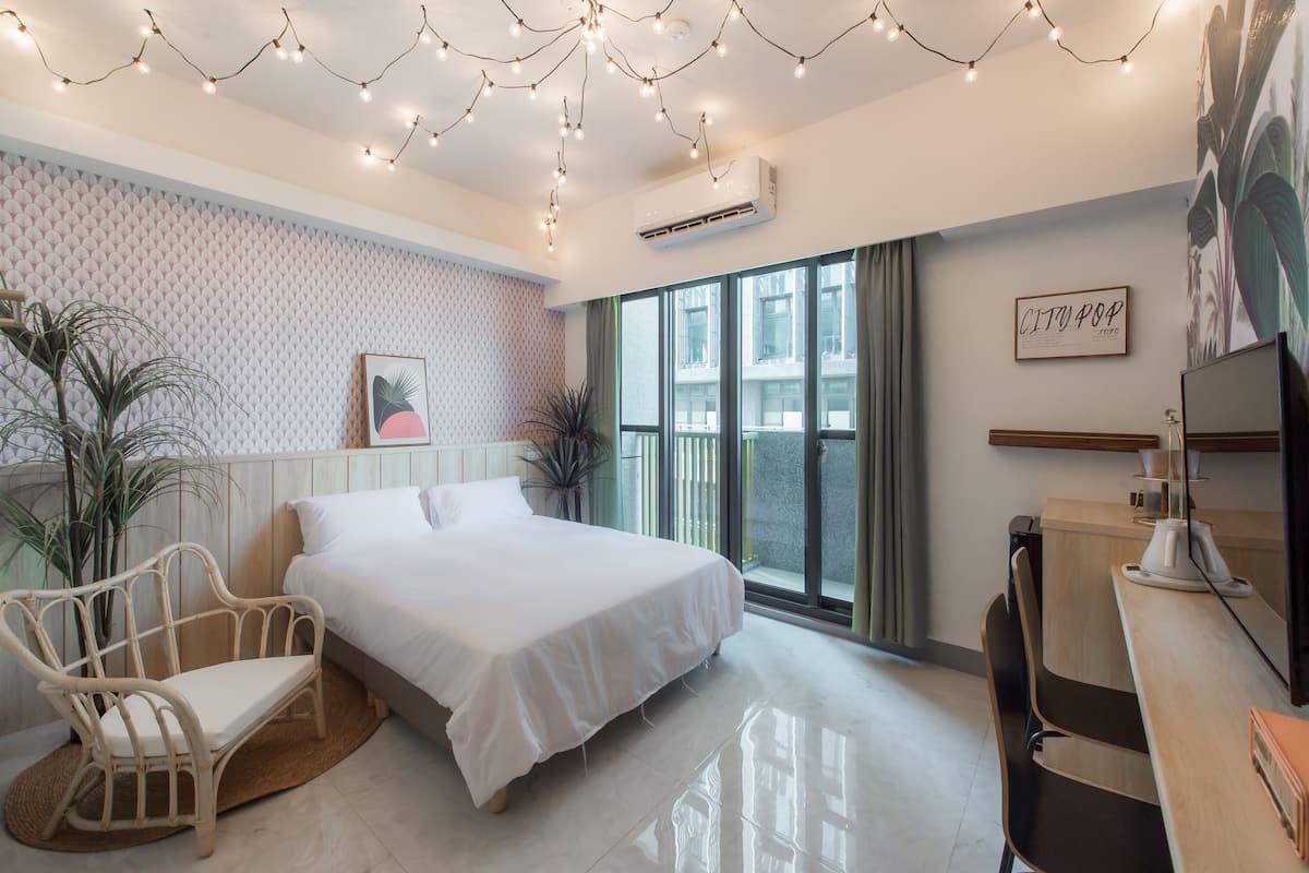 A modern bedroom with a large bed, a desk, and a chair. There are potted plants, string lights, and a window with a view.