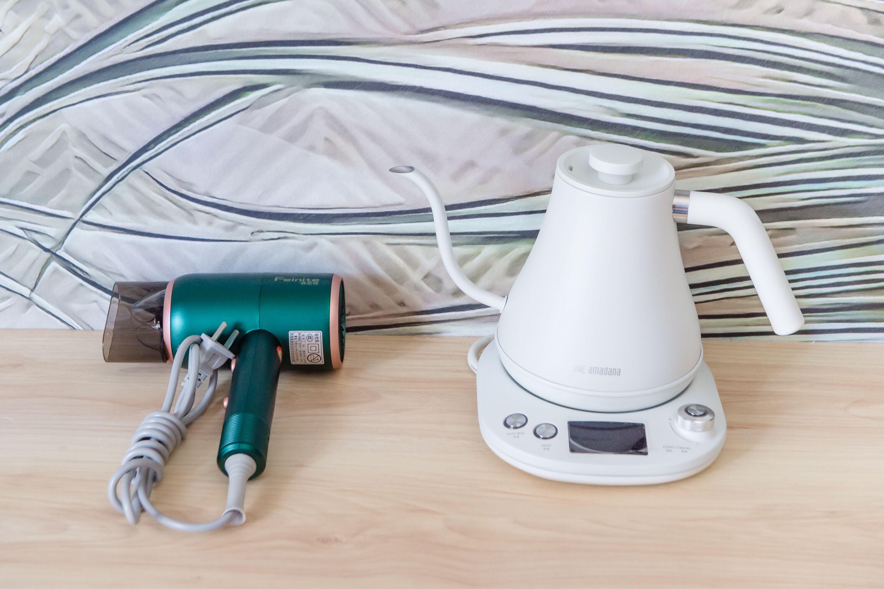 Un assecador i una olla elèctrica sobre una superfície de fusta amb una paret decorada al fons.