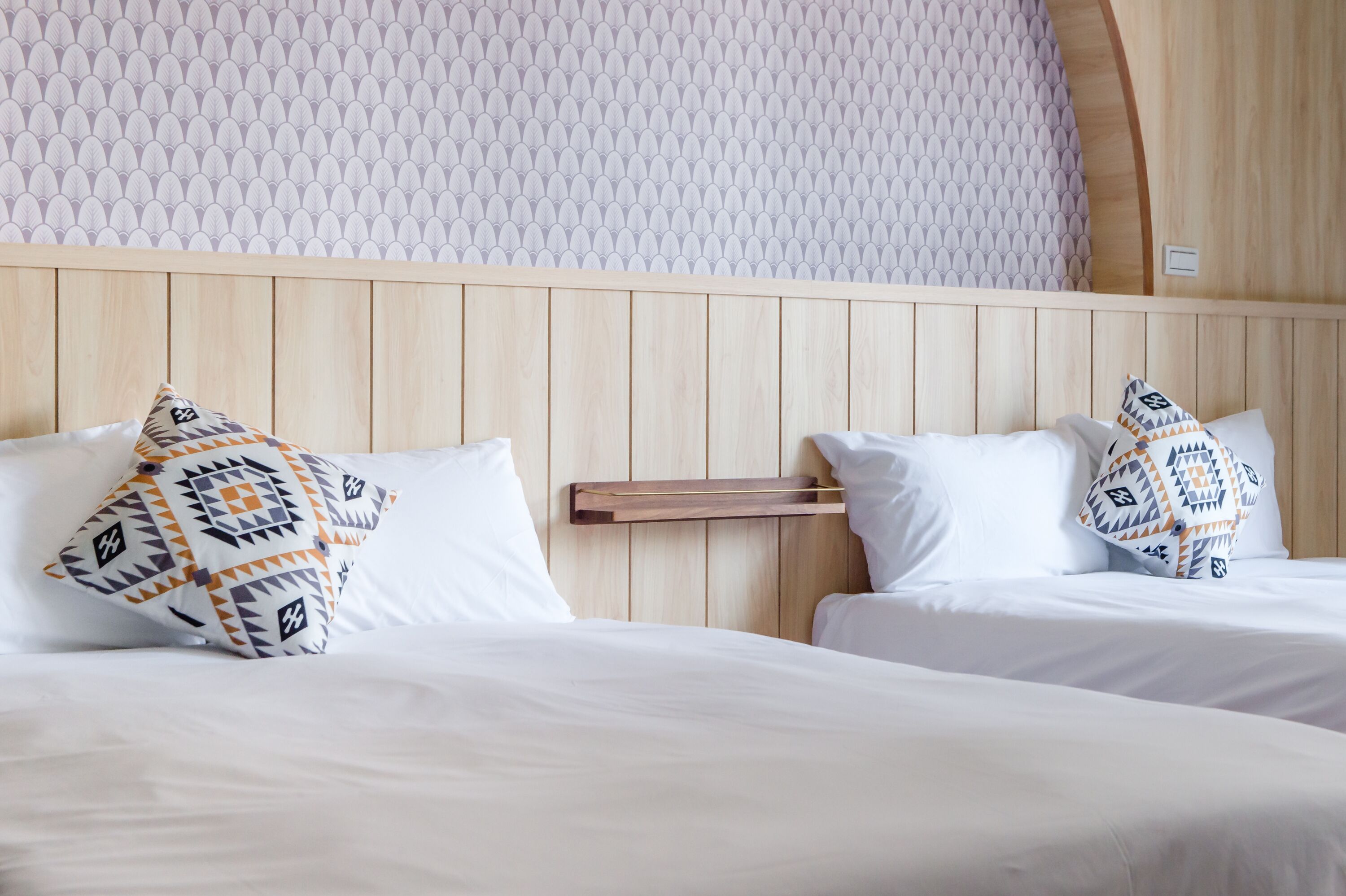 Habitación de hotel con dos camas, cada una con ropa de cama blanca y almohadas estampadas. La habitación tiene un cabecero de madera y papel tapiz con motivos.