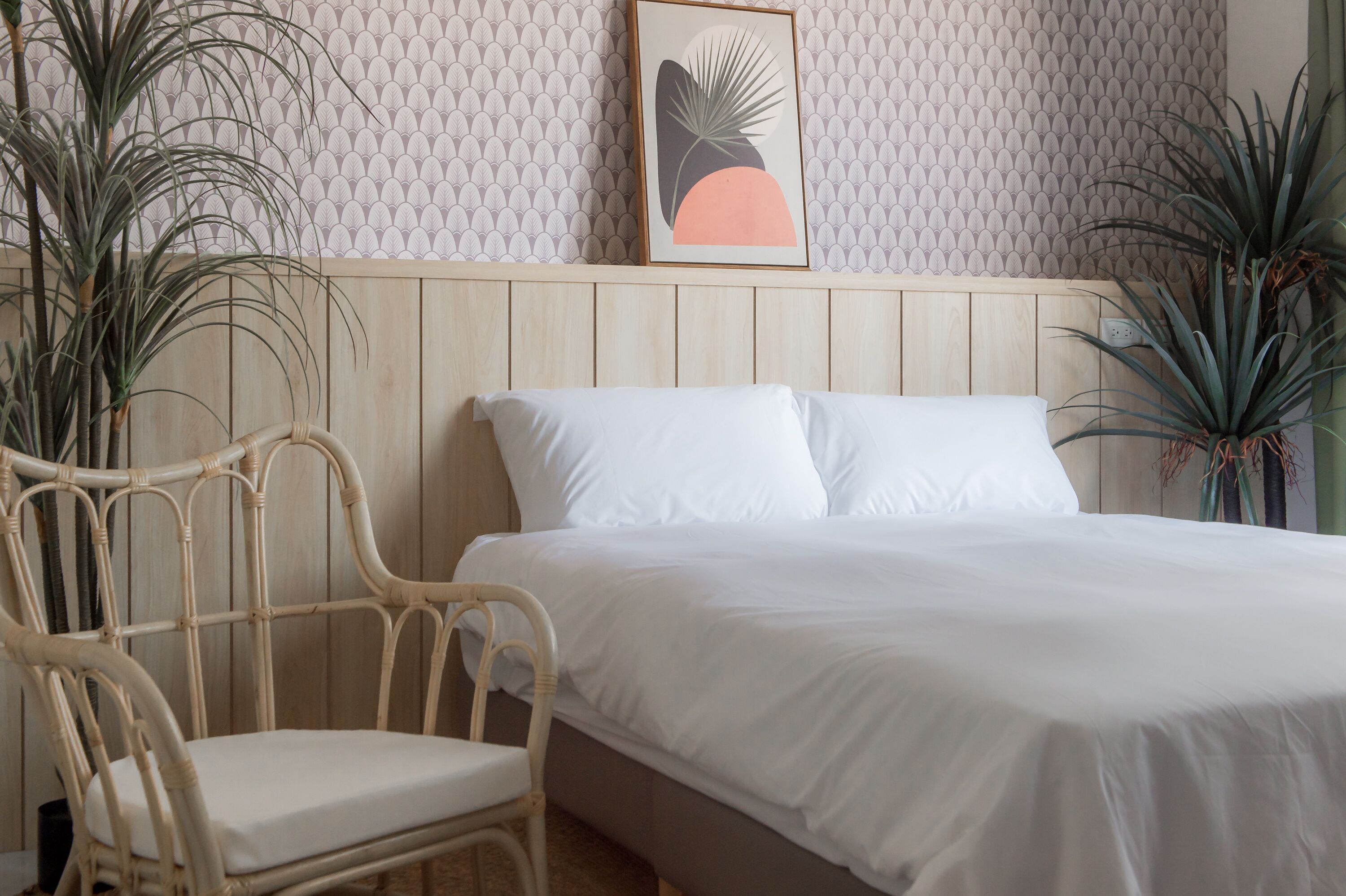 Quarto com cama, duas almofadas brancas, cabeceira de madeira, cadeira de vime e duas plantas em vasos.