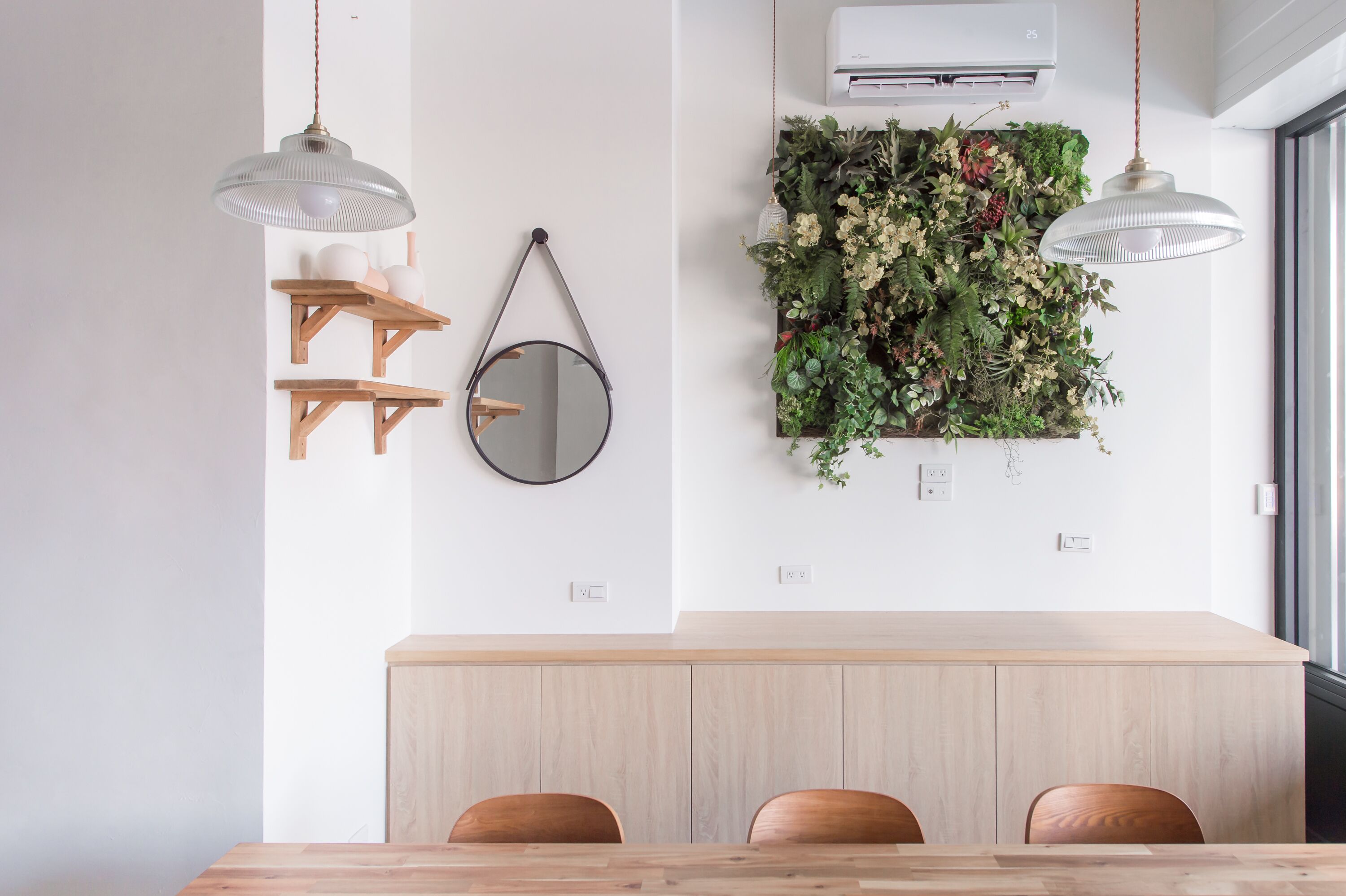 A dining area with a wooden table, two hanging pendant lights, a round mirror, and a wall-mounted air conditioner.