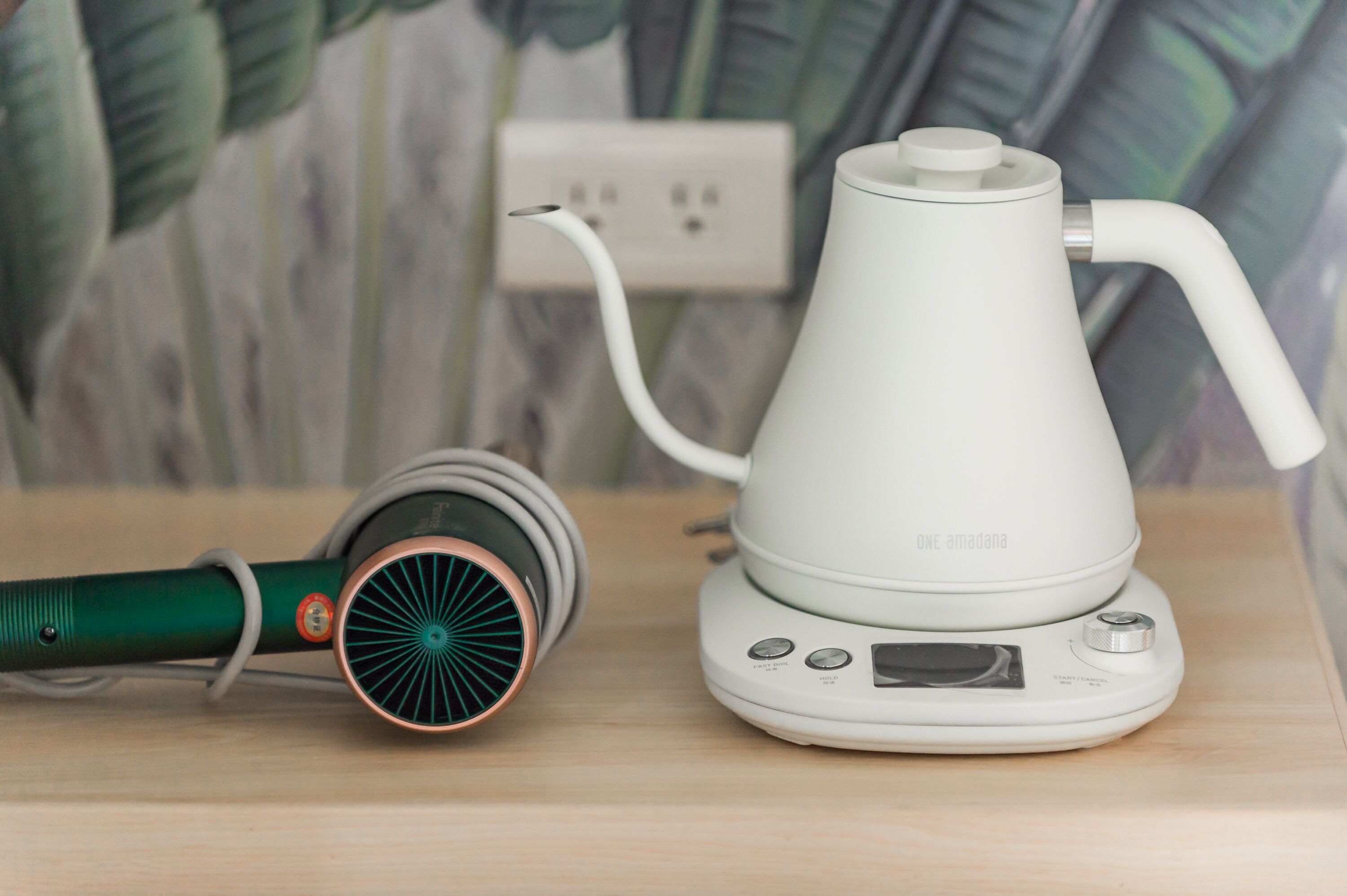 A white electric kettle and a green hairdryer on a wooden surface with a power outlet in the background.