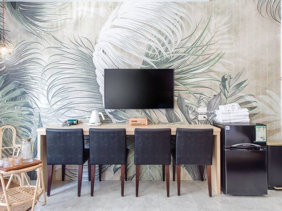 A modern room with a large flat-screen TV mounted on the wall, a wooden desk with three black chairs, a black refrigerator, and a coffee maker.