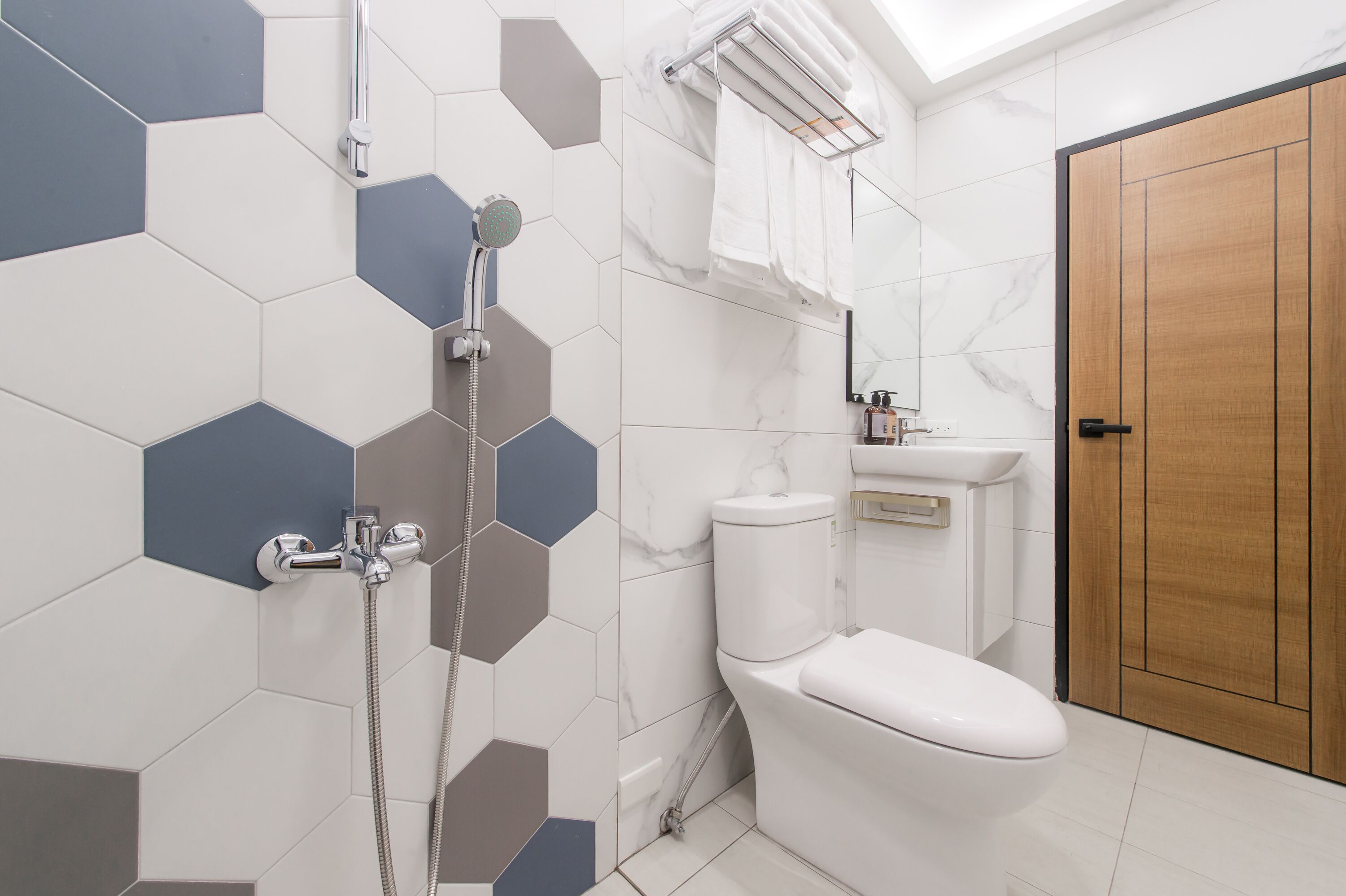 Casa de banho moderna com chuveiro, sanita e lavatório, com azulejos hexagonais e porta de madeira.
