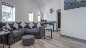 Una sala de estar moderna con un sofá seccional gris, una mesa de centro redonda y un pequeño reposapiés. Se observa una cocina al fondo con una barra y taburetes.