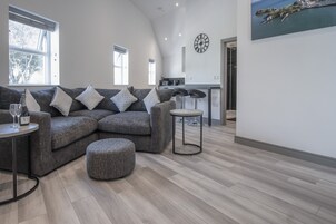 Ein modernes Wohnzimmer mit einem grauen Ecksofa, einem runden Couchtisch und einem kleinen Hocker. Im Hintergrund ist eine Küche mit einer Theke und Hockern zu sehen.