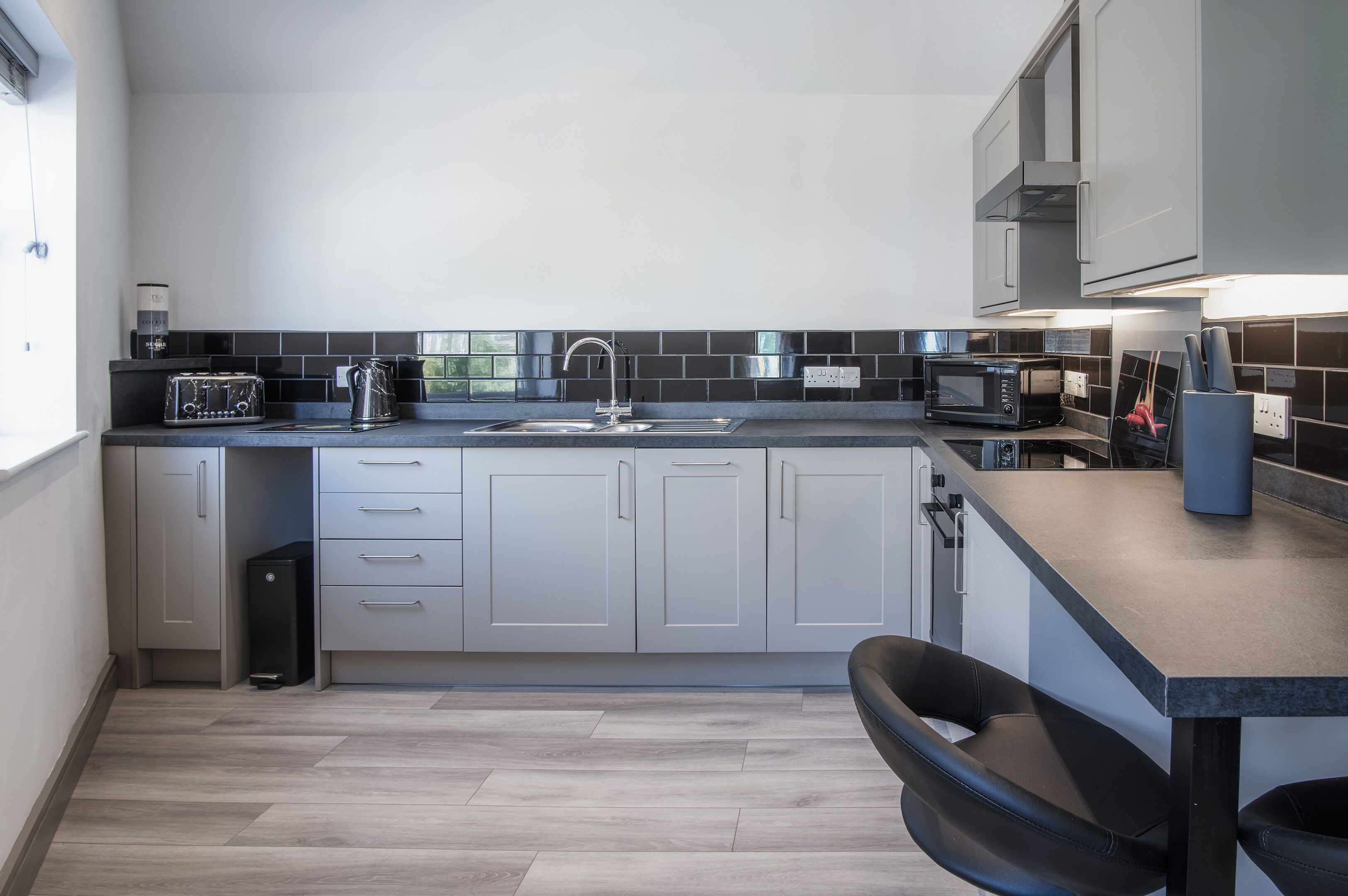 A modern kitchen with grey cabinets, a black countertop, a sink, a kettle, a toaster, a microwave, and a coffee machine.