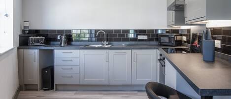 A modern kitchen with grey cabinets, a black countertop, a sink, a kettle, a toaster, a microwave, and a coffee machine.