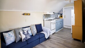 Une cuisine compacte équipée d’une cuisinière, d’un évier et de meubles bleus. Un canapé bleu foncé, agrémenté de coussins à motifs, est placé contre le mur. Un meuble en bois est visible sur la droite.