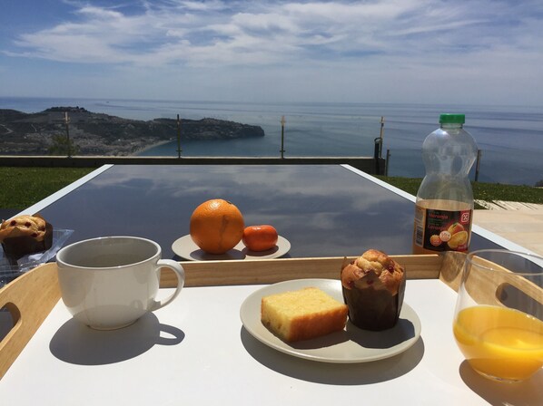 A breakfast tray with a muffin, orange, juice, and a bottle of Naranja Laranja juice on a table with a scenic view.