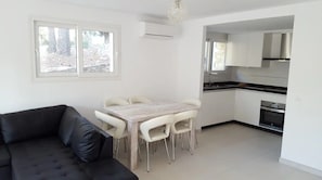 A modern living space with a dining table, chairs, a sofa, a kitchen area, and a window with a view of trees.
