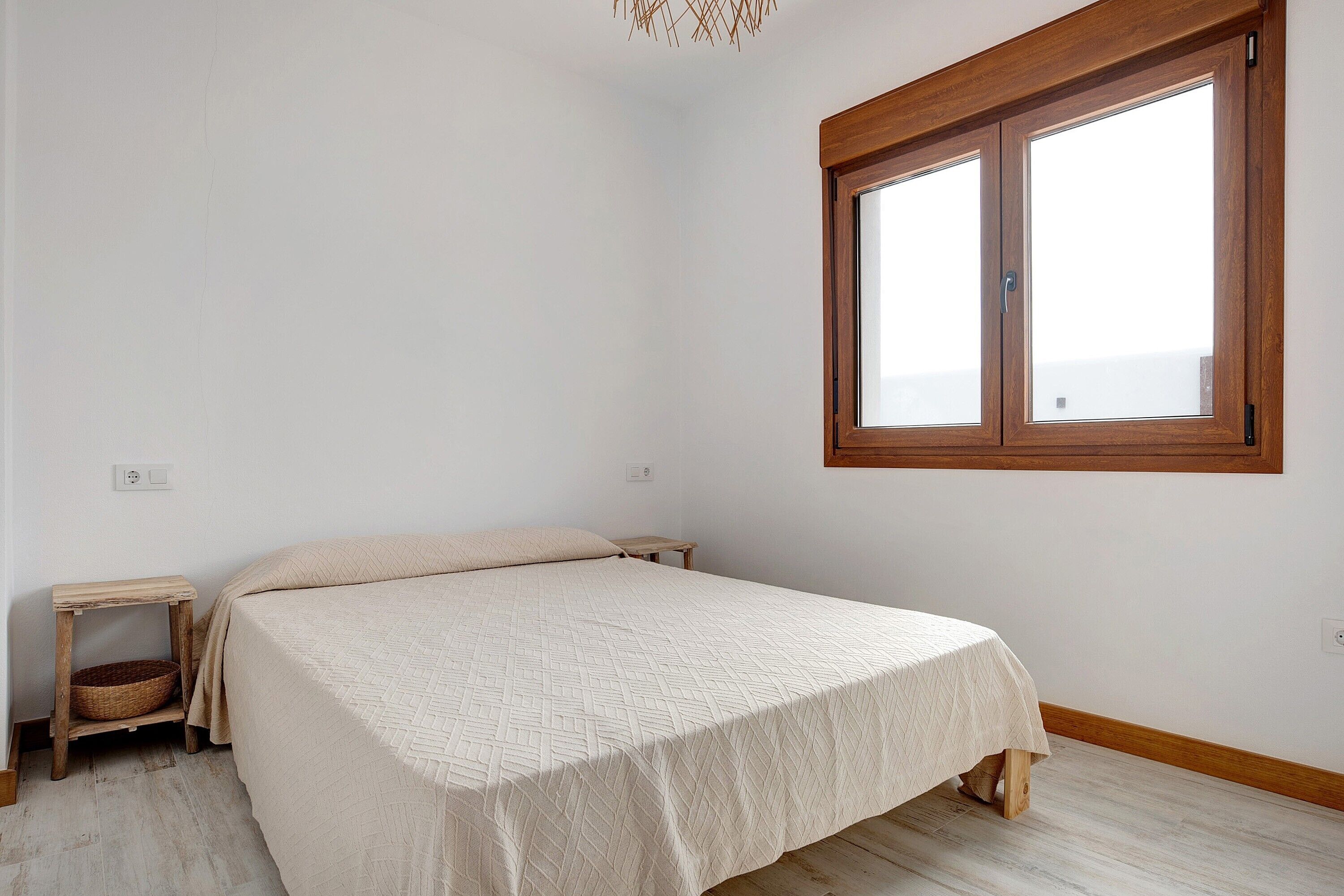 A bedroom with a bed, a small wooden nightstand, a window, and a light switch on the wall.