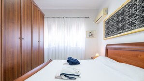 Ein Schlafzimmer mit einem Bett, einem Holzschrank, Vorhängen, einem gerahmten Kunstwerk und einer Klimaanlage.