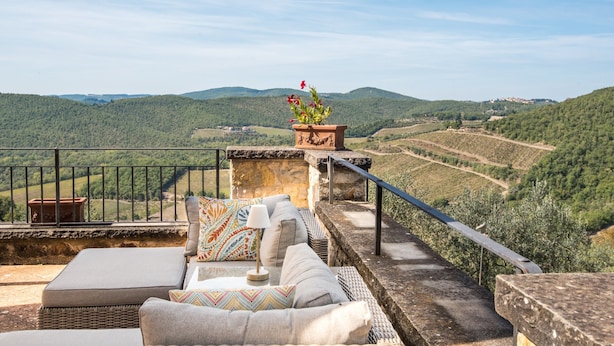Una terraza con mobiliario de mimbre, una maceta de piedra con flores y vistas a viñedos y colinas.