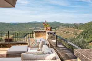 A terrace with wicker furniture, a stone planter with flowers, and a view of vineyards and hills.