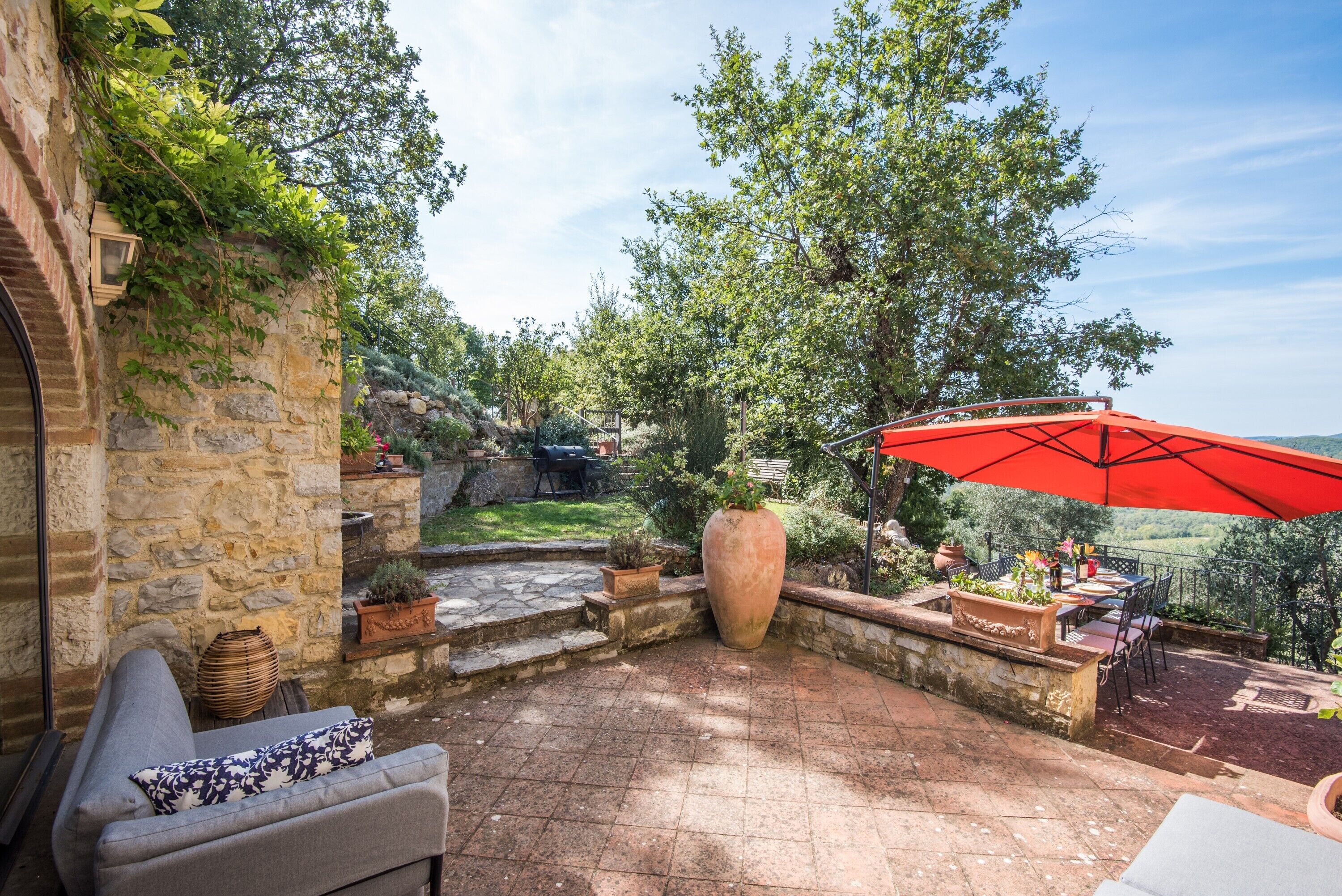 Une terrasse en pierre avec un banc intégré, un grand bac à plantes et un parasol rouge.