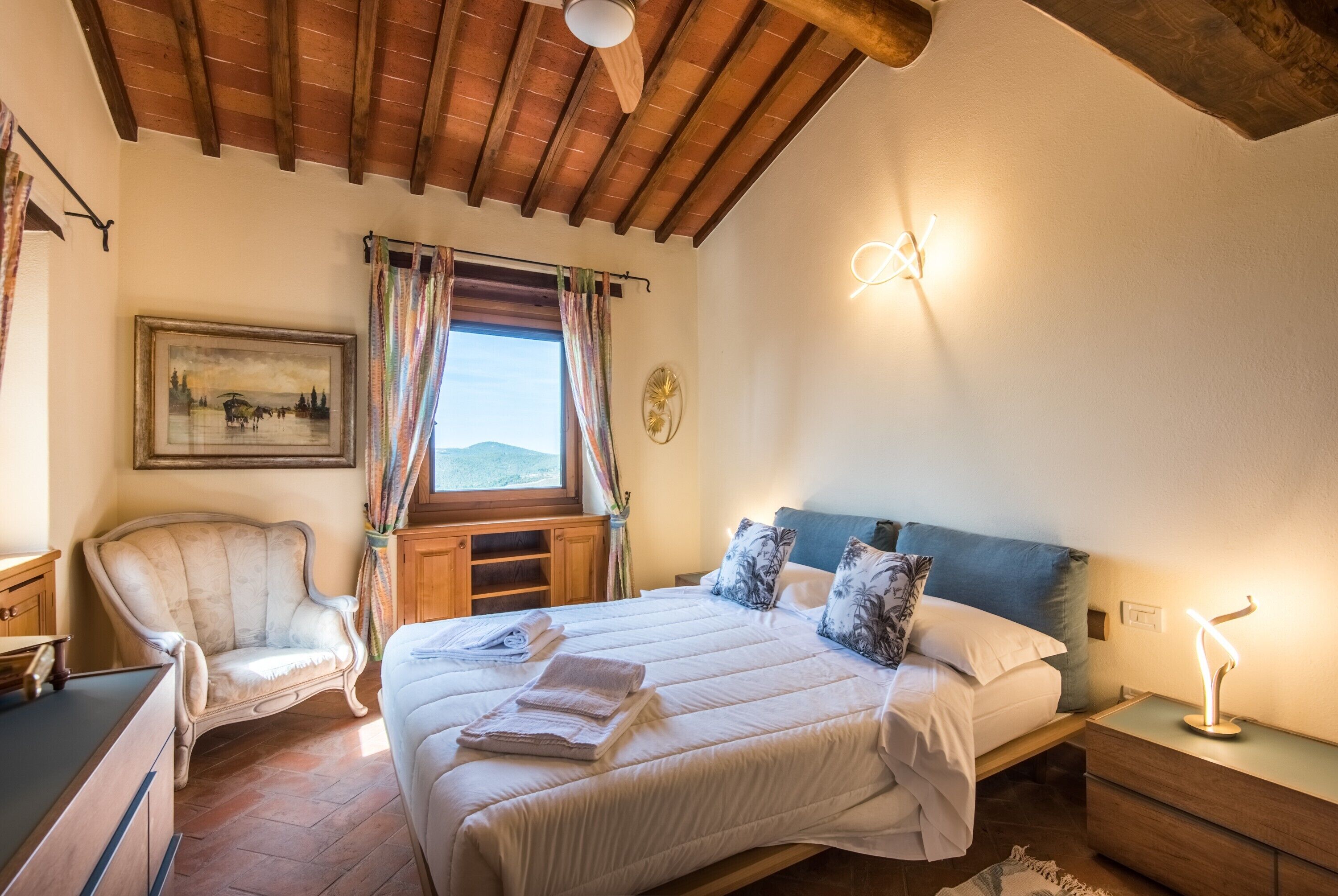 Un dormitorio con una cama, una silla, un escritorio, una ventana con cortinas, un cuadro y una lámpara de pared.