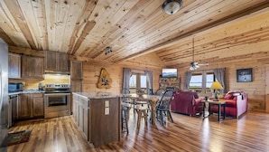 A cabin interior with wooden floors, a kitchen with modern appliances, a dining area with a wooden table and chairs, and a living area with a red sofa and a television.