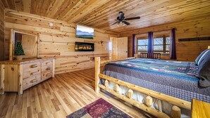 A wooden cabin bedroom with a bed, dresser, TV, fireplace, and windows.