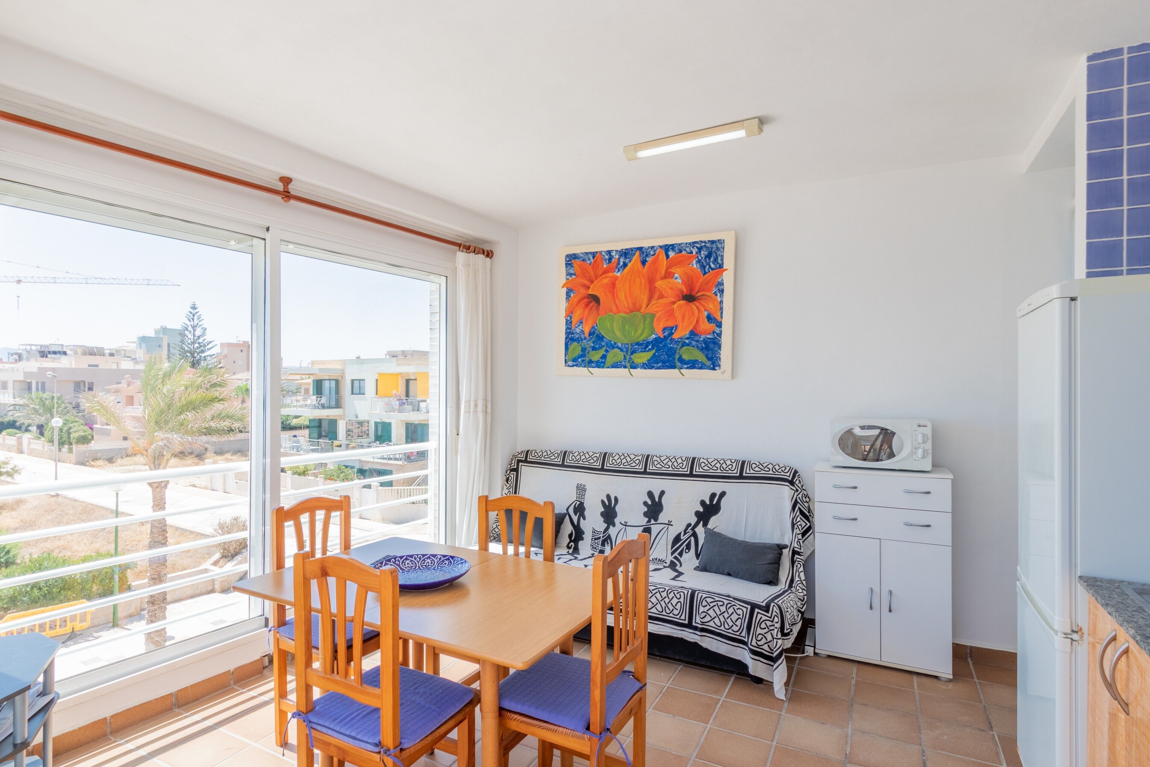 A room with a dining table, chairs, a sofa, a kitchen area, and a large window with a view of buildings.