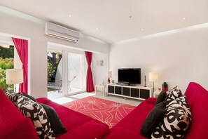 Une chambre d’hôtel moderne avec un canapé rouge, un téléviseur à écran plat, une console blanche et une grande fenêtre ornée de rideaux rouges.