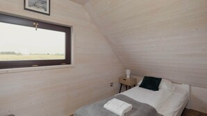 A bedroom with a wooden sloped ceiling, a window, a bedside table, and a bed with a white mattress and gray blanket. - Apartment with Garden | Parking, 4.9 km to the Beach (Powiat koszaliński)