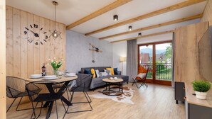 A modern living room with a wooden ceiling, a dining area with black chairs, a grey sofa with colorful cushions, and a large window with a view of the outdoors. - Atmospheric Apartment | Mountain View and Workspace (Zakopane)