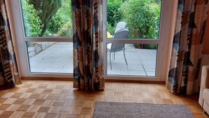 Ein Zimmer mit einer großen Schiebeglasdoor zur Terrasse. Der Boden ist mit einem Fischgrätmuster aus Holz versehen und mit einem grauen Webteppich ausgelegt.