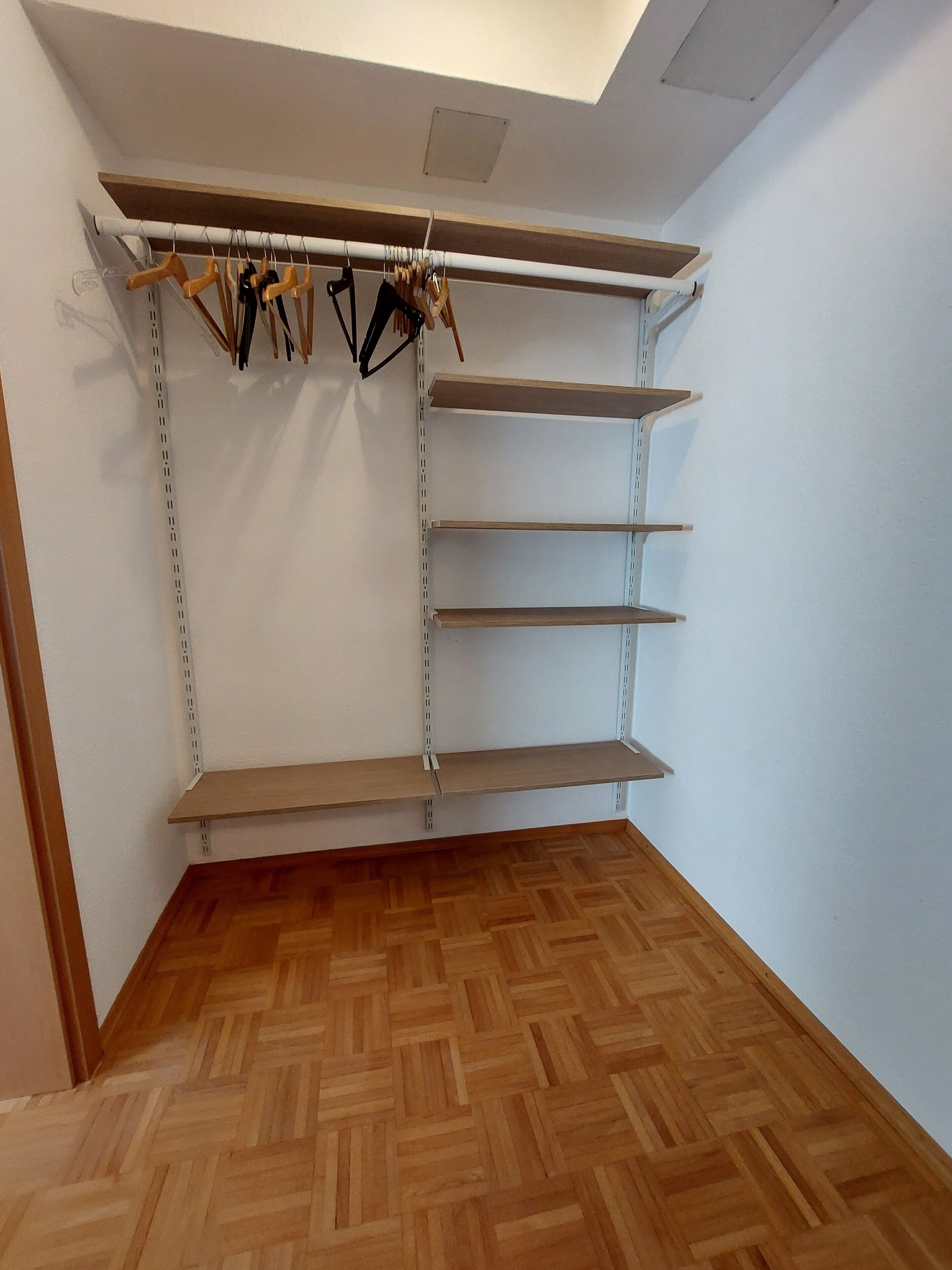 Un coin de pièce avec un parquet en bois disposé en chevrons, un mur blanc et une étagère en métal avec des étagères en bois.