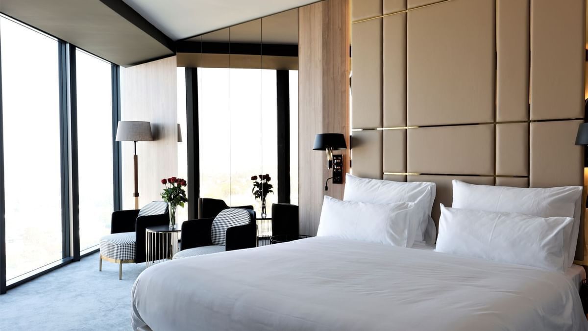 Una cama grande con sábanas blancas, una mesita de noche, una lámpara y una silla en una habitación de hotel moderna con ventanas amplias.